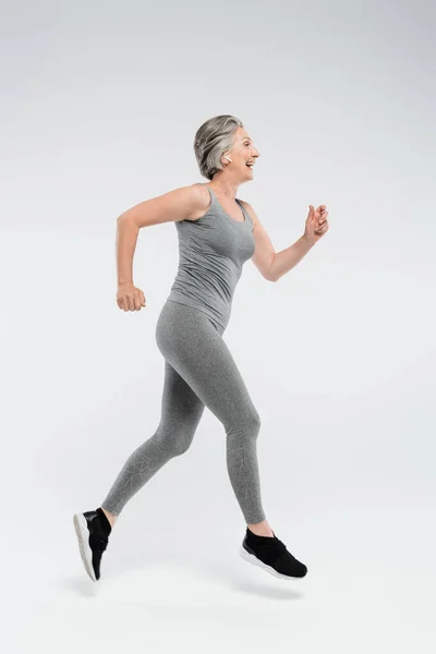 Full length of happy senior woman with grey hair running on grey background — Stock Photo