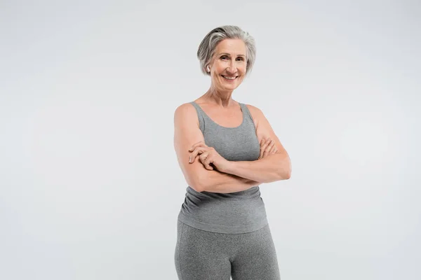 Веселая пожилая женщина в спортивной одежде стоящая со сложенными руками изолированная на сером — стоковое фото