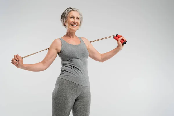 Cheerful senior woman with grey hair holding skipping rope isolated on grey — Stock Photo