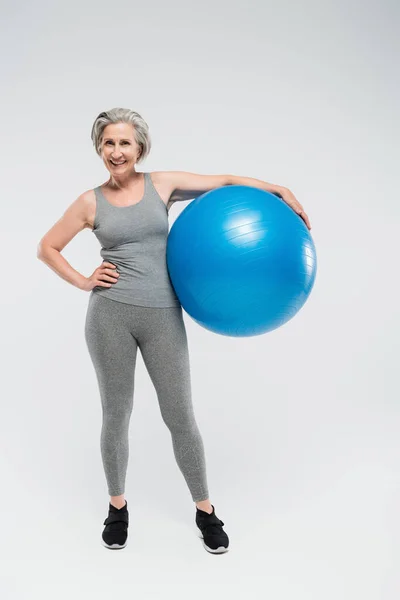 Piena lunghezza di felice donna anziana in abbigliamento sportivo tenendo palla fitness su grigio — Foto stock