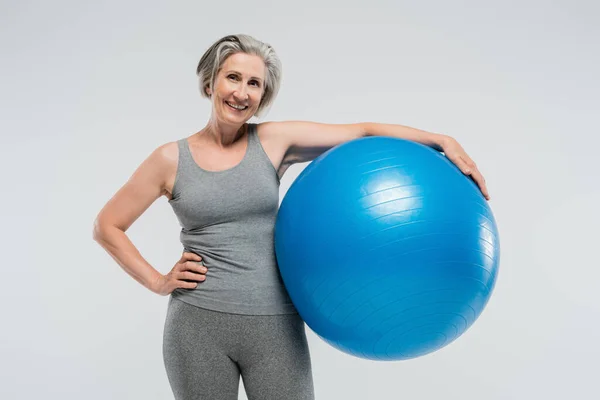 Весела старша жінка в спортивному одязі тримає синій фітнес-куля ізольована на сірому — стокове фото