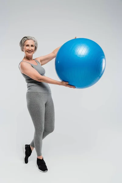 Pleine longueur de femme âgée gaie en vêtements de sport tenant ballon de fitness bleu sur gris — Photo de stock