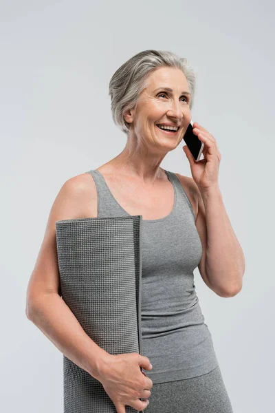 Happy grandma holding fitness mat and talking on mobile phone isolated on grey — Stock Photo