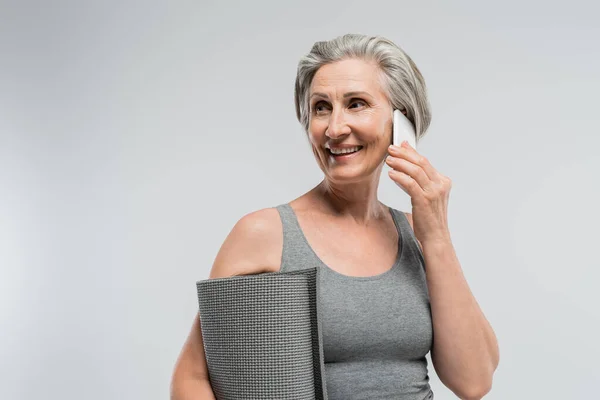 Überglückliche Oma hält Fitnessmatte in der Hand und telefoniert isoliert auf grau — Stockfoto