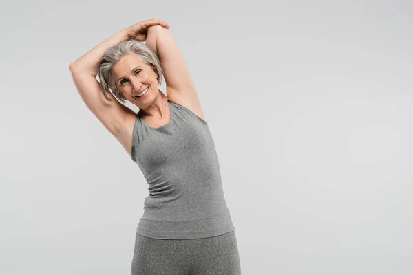 Жизнерадостная бабушка в спортивной одежде, упражняющаяся и улыбающаяся изолированная на сером — стоковое фото