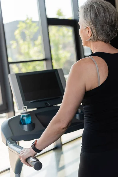 Senior woman with grey hair running on treadmill next to sports bottle with water — Stock Photo
