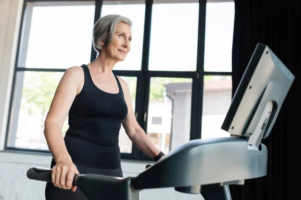 Gaie femme âgée avec les cheveux gris courir sur tapis roulant dans la salle de gym — Photo de stock