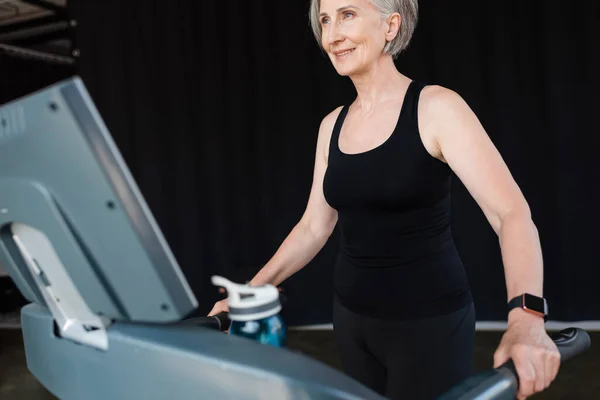 Mujer mayor positiva con pelo gris haciendo ejercicio en la cinta de correr en el gimnasio - foto de stock