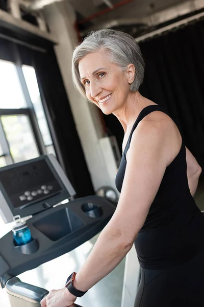Happy senior woman with grey hair running on treadmill next to sports bottle with water — Stock Photo