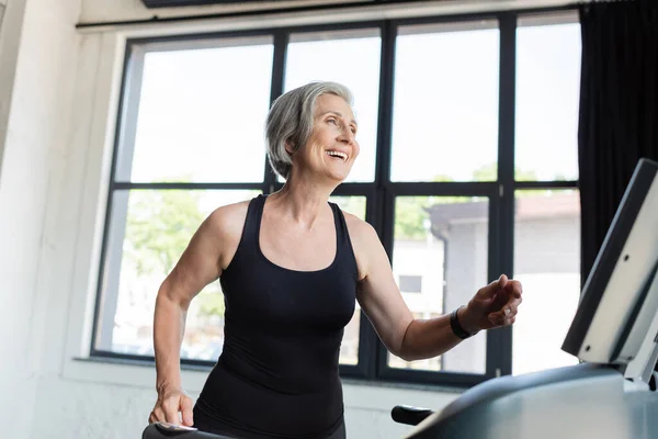 Mujer mayor muy alegre con el pelo gris corriendo en la cinta de correr en el gimnasio - foto de stock