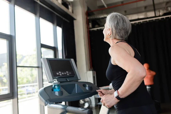 Retired woman with grey hair running on treadmill next to sports bottle with water — Stock Photo