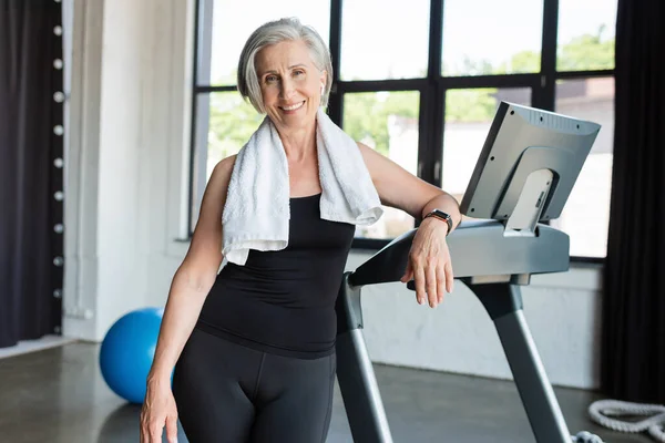 Happy senior woman with white towel on shoulders standing next to treadmill — Stock Photo