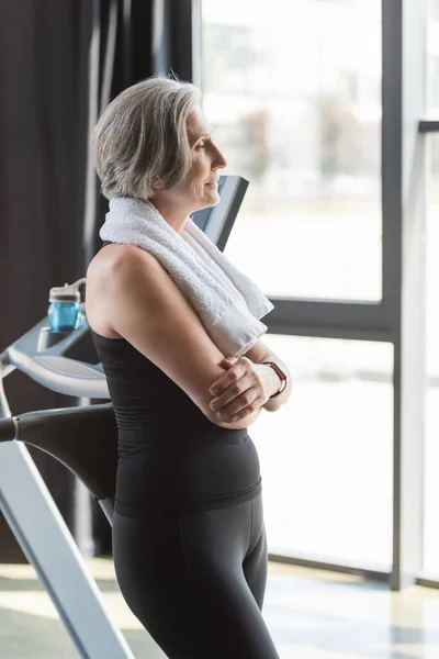 Happy senior woman with white towel on shoulders standing with crossed arms next to treadmill — Stock Photo