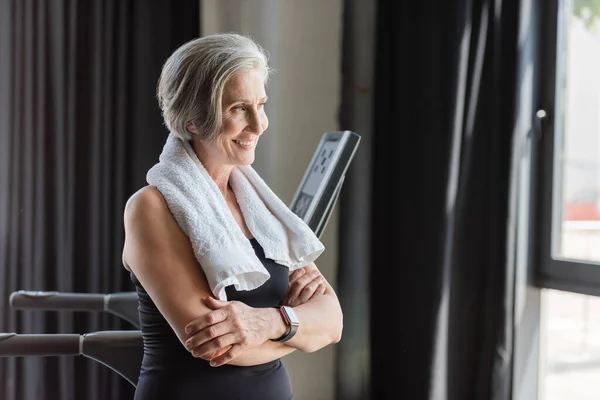 Happy senior woman with white towel on shoulders standing with folded arms next to treadmill — Stock Photo