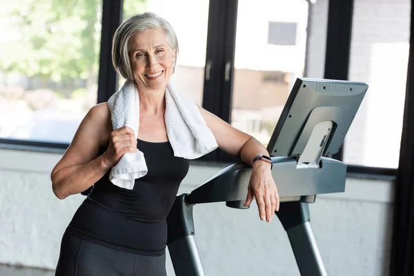 Joyful retired woman with white towel on shoulders standing next to treadmill — Stock Photo