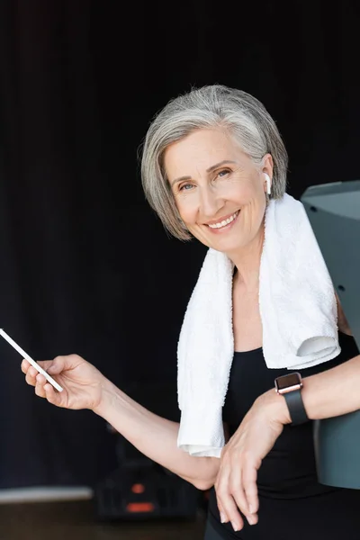 Mujer alegre y mayor en auriculares inalámbricos con teléfono inteligente en el gimnasio - foto de stock