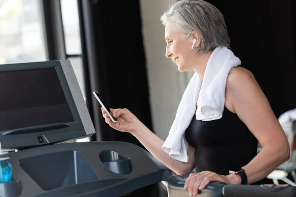 Happy retired woman in wireless earphone holding smartphone while standing near treadmill in gym — Stock Photo