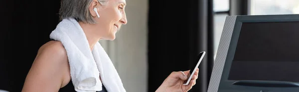 Heureuse femme âgée dans un écouteur sans fil tenant smartphone tout en se tenant près du tapis roulant dans la salle de gym, bannière — Photo de stock