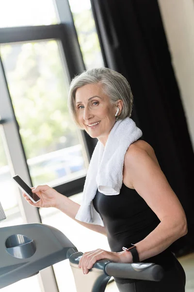 Joyful senior woman in wireless earphone holding smartphone while standing near treadmill in gym — Stock Photo