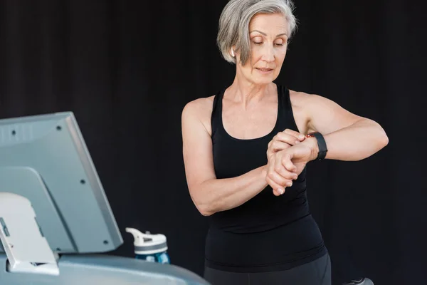 Seniorin mit grauen Haaren checkt Aktivität auf Fitness-Tracker nach Ausdauertraining auf Laufband — Stockfoto