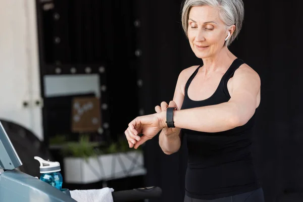 Mujer mayor satisfecha con la actividad de verificación de auriculares inalámbricos en el rastreador de fitness en el gimnasio - foto de stock