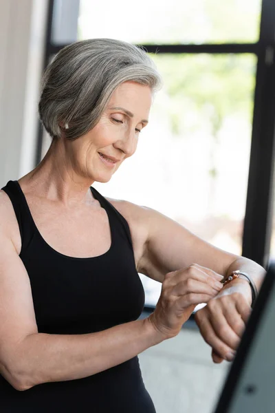 Heureuse femme âgée avec une activité de vérification des cheveux gris sur le traqueur de fitness dans la salle de gym — Photo de stock
