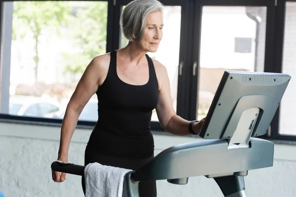 Старша жінка з сірим волоссям дивиться на монітор бігової доріжки під час роботи в спортзалі — стокове фото