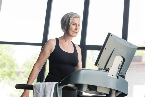 Retired woman with grey hair looking at monitor of treadmill while working out in gym — Stock Photo