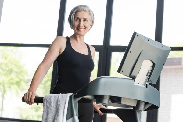 Mujer mayor feliz con el pelo gris mirando a la cámara mientras hace ejercicio en el gimnasio - foto de stock