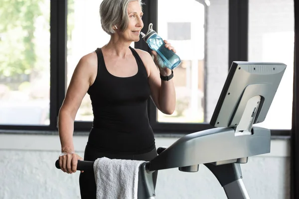 Seniorin mit grauen Haaren trinkt nach Ausdauertraining auf Laufband Wasser aus Sportflasche — Stockfoto