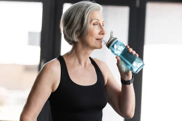 Donna anziana con capelli grigi che beve acqua dalla bottiglia sportiva in palestra — Foto stock