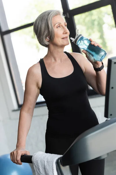 Mujer jubilada con el pelo gris beber agua de la botella de deportes después de ejercicio cardiovascular en la cinta de correr - foto de stock