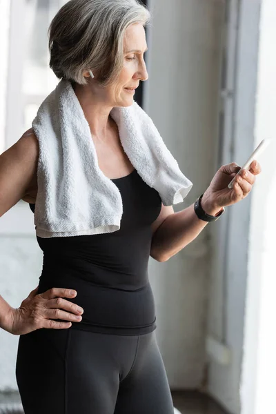 Mujer mayor en forma con toalla en los hombros usando un teléfono inteligente mientras escucha música en auriculares inalámbricos - foto de stock