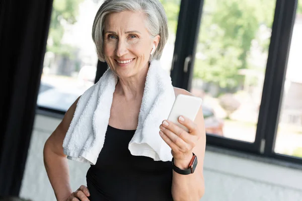 Fröhliche Seniorin mit Handtuch auf den Schultern und Smartphone beim Musikhören im drahtlosen Kopfhörer — Stockfoto