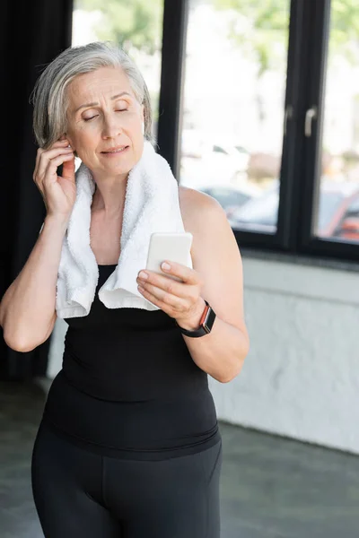 Fit senior woman with towel on shoulders using smartphone while wearing wireless earphone — Stock Photo