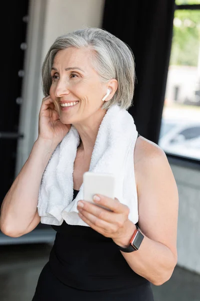 Mujer mayor feliz con toalla en los hombros usando un teléfono inteligente mientras usa auriculares inalámbricos - foto de stock