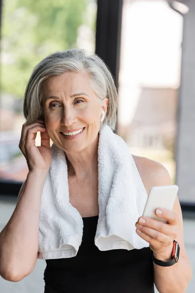 Felice donna anziana con asciugamano sulle spalle utilizzando smartphone e regolazione auricolare wireless — Foto stock