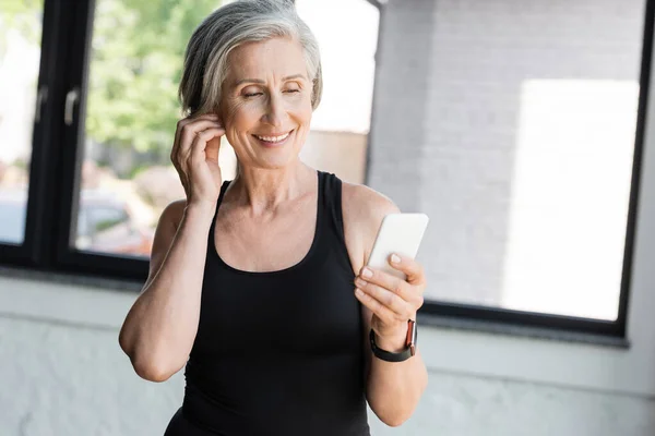 Caben mujer mayor en camiseta negra usando teléfono inteligente mientras escucha música en auriculares inalámbricos - foto de stock