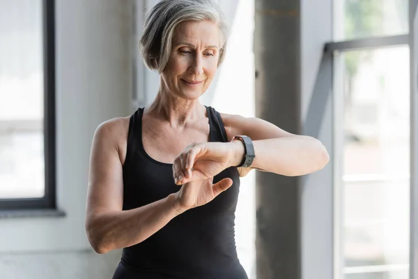 Happy senior woman in black tank top checking activity on fitness tracker in gym — Stock Photo