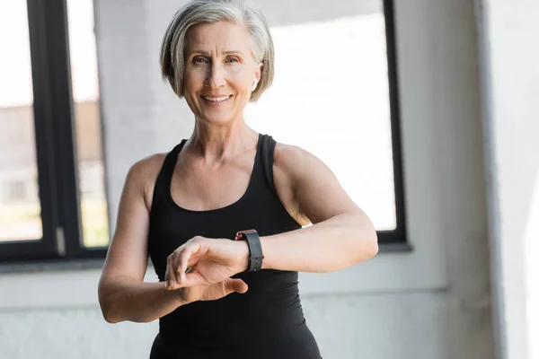 Fit senior femme en noir débardeur souriant tout en vérifiant l'activité sur fitness tracker dans la salle de gym — Photo de stock