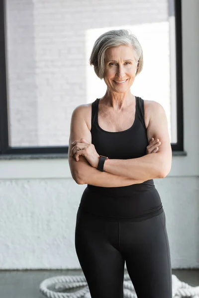 Fitte Seniorin in schwarzem Tank-Top lächelnd im Stehen mit verschränkten Armen im Fitnessstudio — Stockfoto