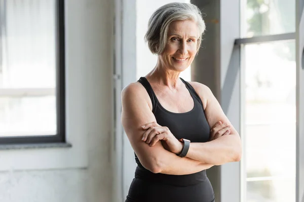 Fit and retired woman in tank top smiling while standing with crossed arms in sports center — Stock Photo