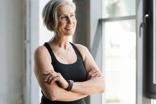 Happy senior sportswoman in tank top smiling while standing with crossed arms in gym — Stock Photo