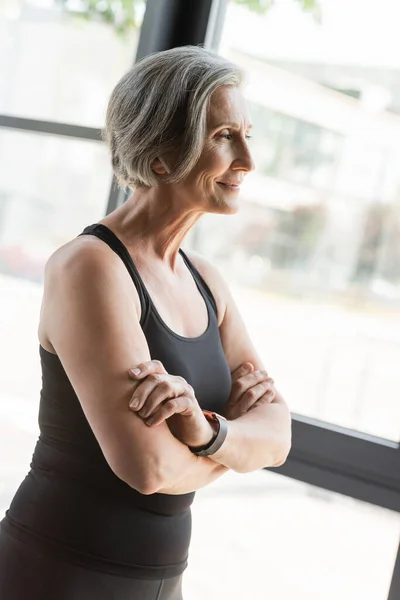 Cheerful senior sportswoman in tank top smiling while standing with crossed arms in gym — Stock Photo