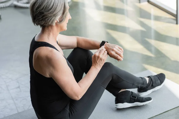 Fit senior woman with grey hair sitting and using fitness tracker after working out in gym — Stock Photo