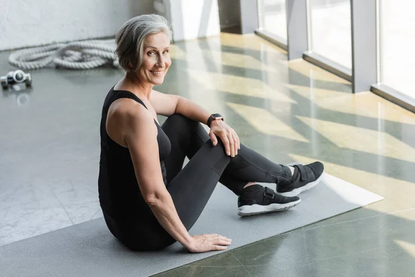 Volle Länge der glücklichen Senior-Sportlerin mit grauen Haaren sitzt auf Fitnessmatte in der Turnhalle — Stockfoto