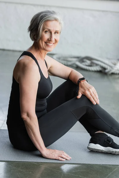 Happy senior sportswoman with grey hair smiling while sitting on fitness mat in gym — Stock Photo