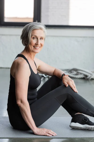 Joyful senior sportswoman with grey hair smiling while sitting on fitness mat in gym — Stock Photo