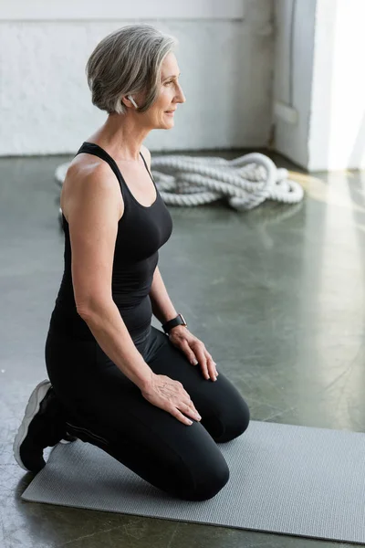 Mujer en forma jubilada escuchando música en los auriculares mientras hace ejercicio en la alfombra de fitness - foto de stock