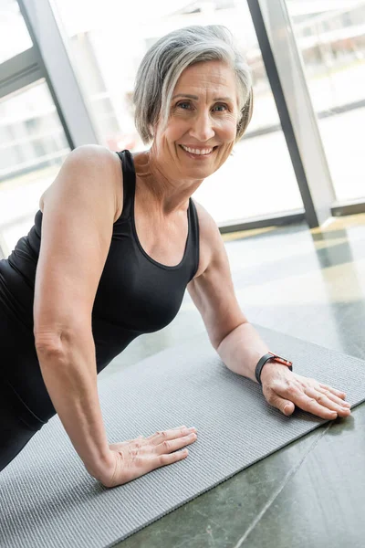 Sportiva anziana sorridente mentre si esercita sul tappetino fitness in palestra — Foto stock